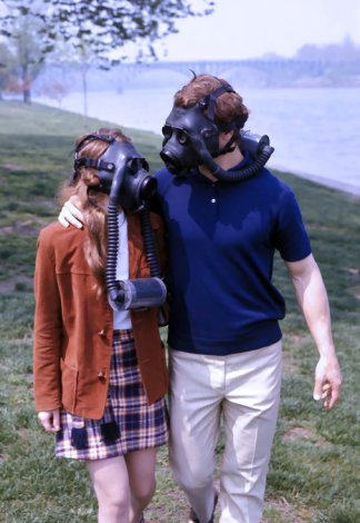 Man with arm around woman walking along grassy river bank, both wearing gas masks